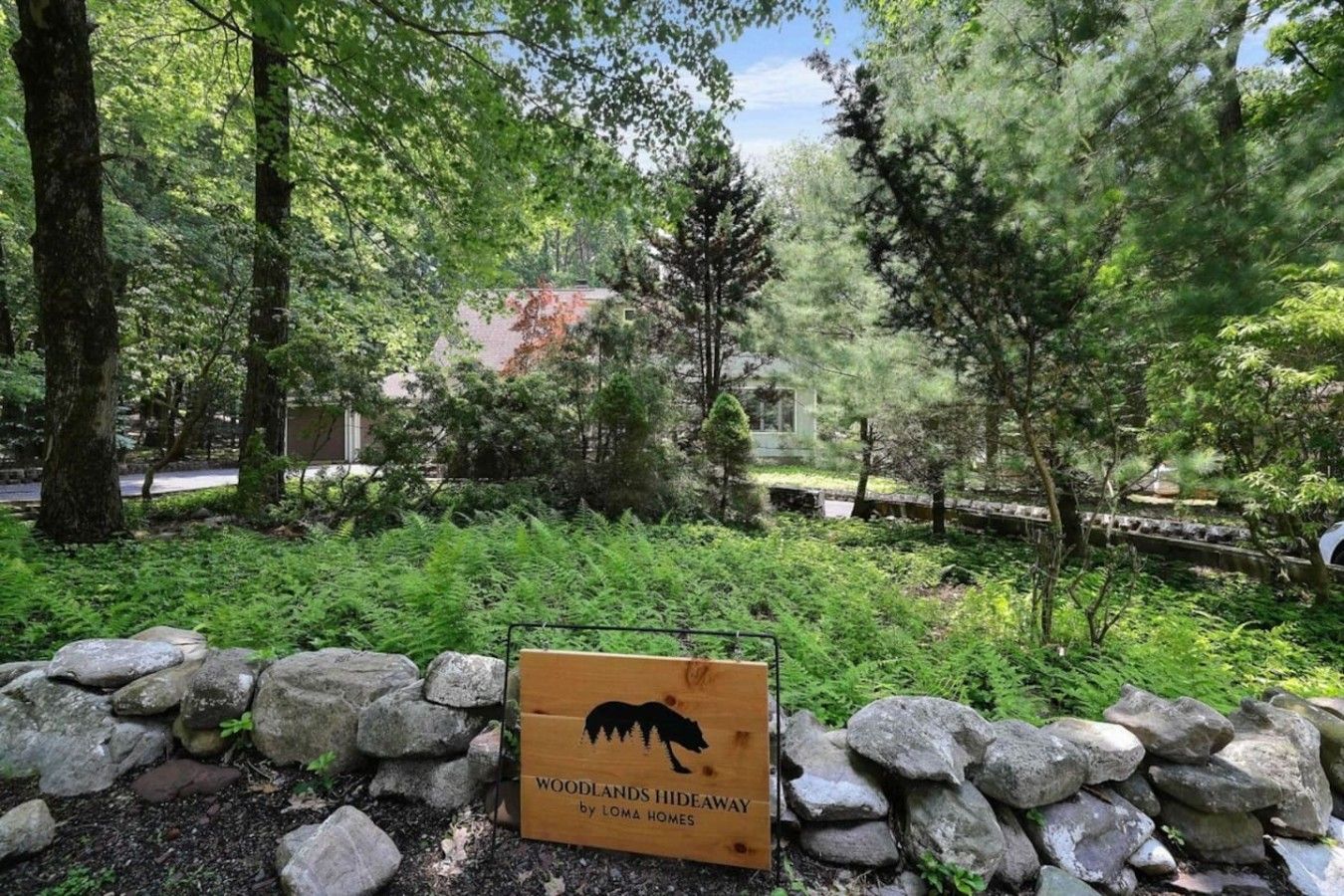 A sign for 'Woodlands Hideaway by Loma Homes' surrounded by a stone wall and dense greenery.