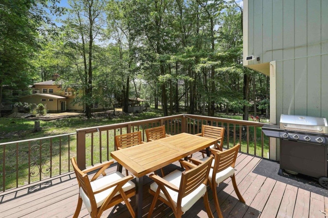 A wooden deck with a table and chairs, a barbecue grill, and a view of a forested area.