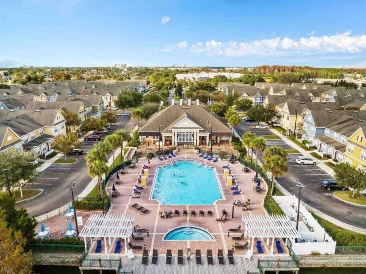 A residential area with a central swimming pool surrounded by lounge chairs and umbrellas, with houses in the background.