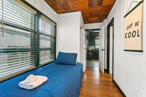 Ein Schlafzimmer mit einem Bett, einem Fenster mit Jalousien, einer Holzbalkendecke und einem Flur, der zu einer Küche führt.