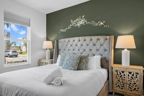 Een netjes opgemaakt bed met een witte deken en kussens, aan weerszijden van twee nachtkastjes met lampen, in een kamer met een groene accentmuur en een raam met uitzicht op een straat.