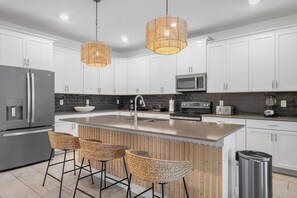 A modern kitchen with white cabinets, a stainless steel refrigerator, a microwave, and a large island with bar stools. - 6BR Champions Gate Villa | w/Oasis Club Amenities! (Davenport)