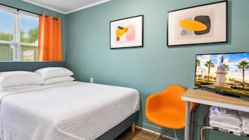 A hotel room with a bed, a desk with a television, two framed artworks, and a window with curtains.