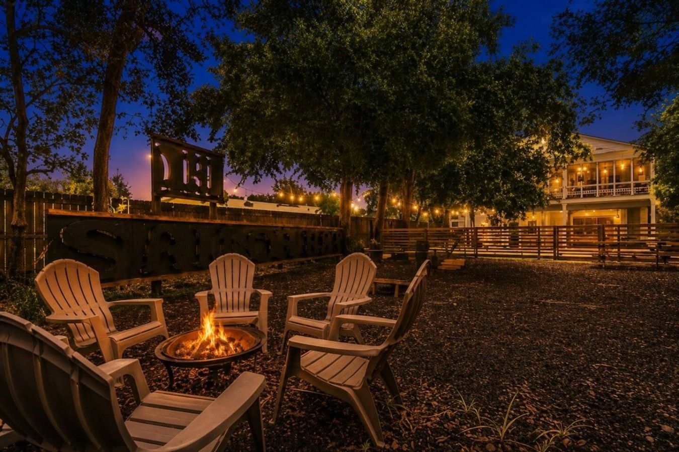 An outdoor seating area with Adirondack chairs and a fire pit, surrounded by trees and a building with string lights.