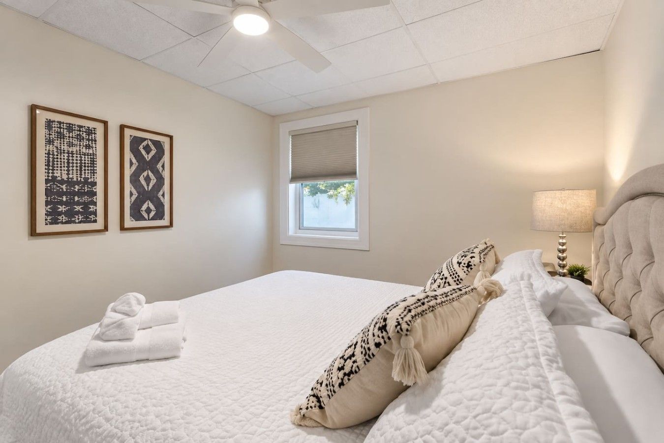 A hotel room with a bed, a ceiling fan, a window with blinds, and two framed wall art pieces.