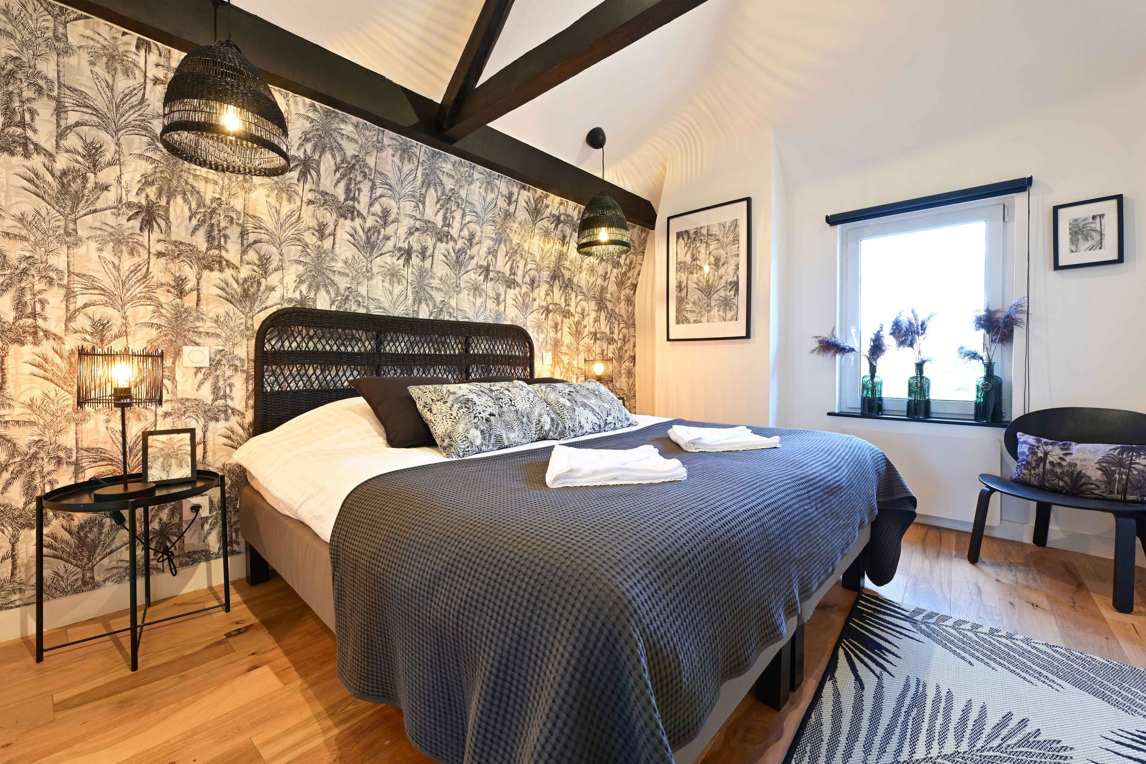 A bedroom with a bed, a nightstand, a window with a view, and a wall with a tropical wallpaper.