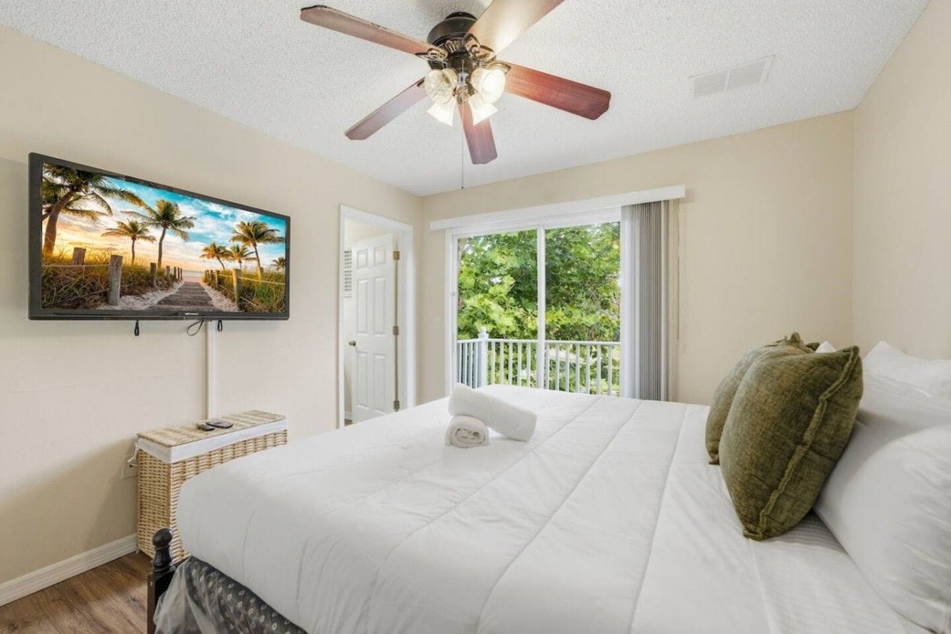 A bedroom with a bed, a ceiling fan, a television on the wall, and a view of greenery outside.