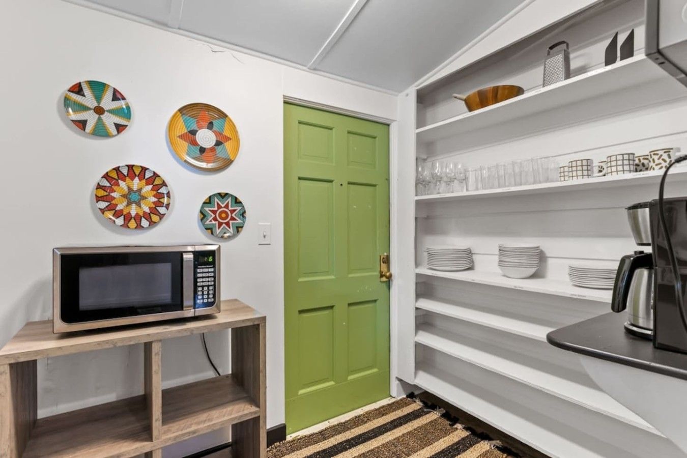 A kitchen with a microwave on a wooden stand, a green door, and shelves with plates and glasses.