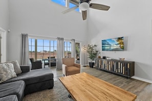 A spacious living room with a ceiling fan, a large window with curtains, a sofa, a coffee table, and a painting on the wall.