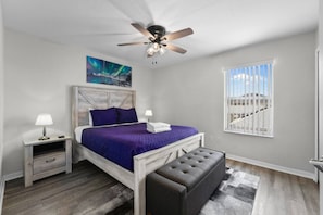 A bedroom with a bed, nightstands, a ceiling fan, a window with a view, and a painting on the wall.