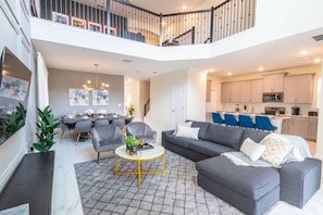 Ein großzügiger Wohnbereich mit grauem Sofa, Sesseln und einem Couchtisch. Im Hintergrund ist ein Essbereich mit Tisch und Stühlen zu sehen.