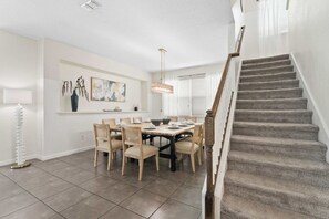 A dining area with a table set for six, a staircase, and a wall with a painting and a vase. - SUPER DREAM – Luxury Poolside Loft | Pool & Games (Kissimmee)
