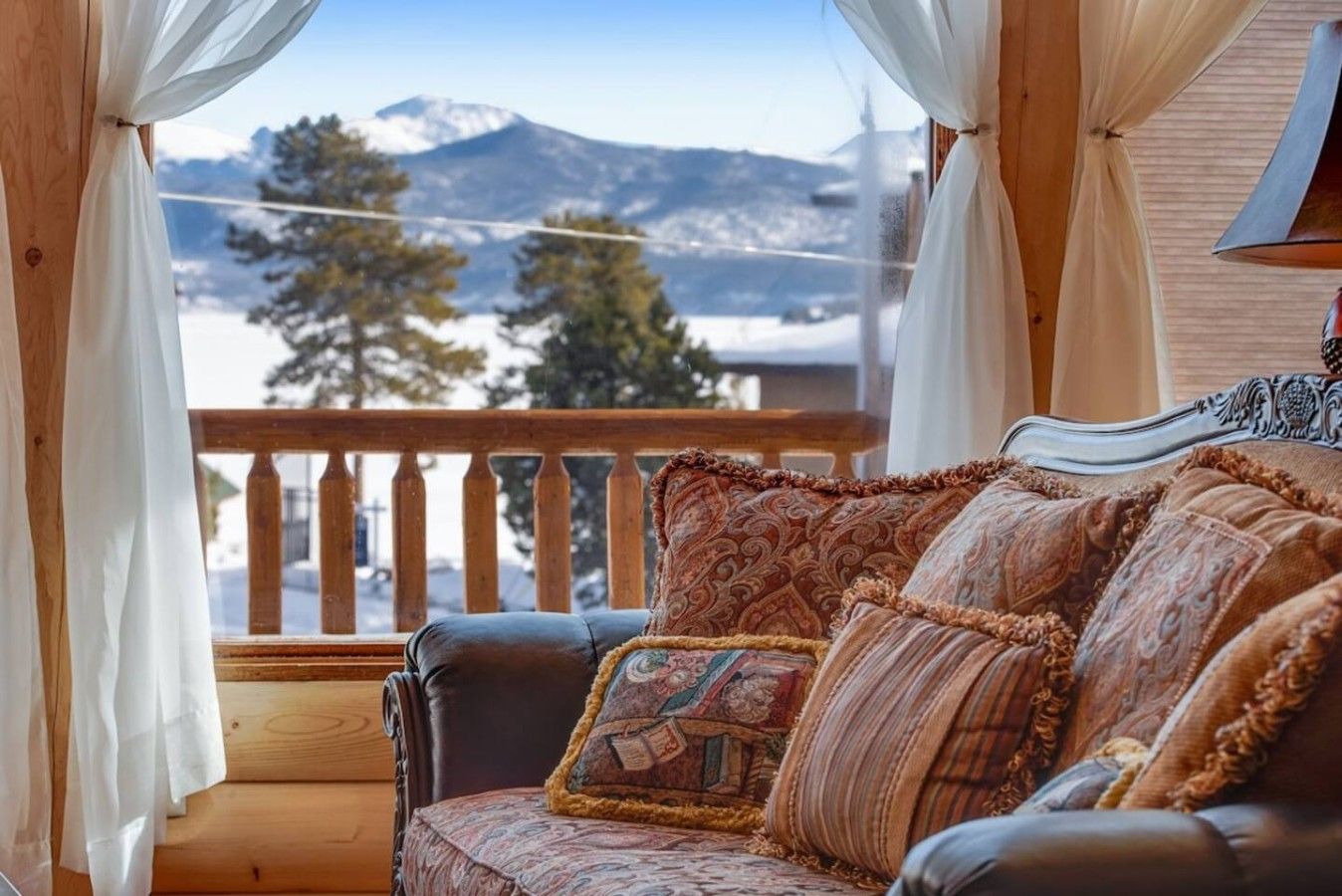 A cozy room with a wooden balcony, a cushioned sofa, and a view of snow-capped mountains.