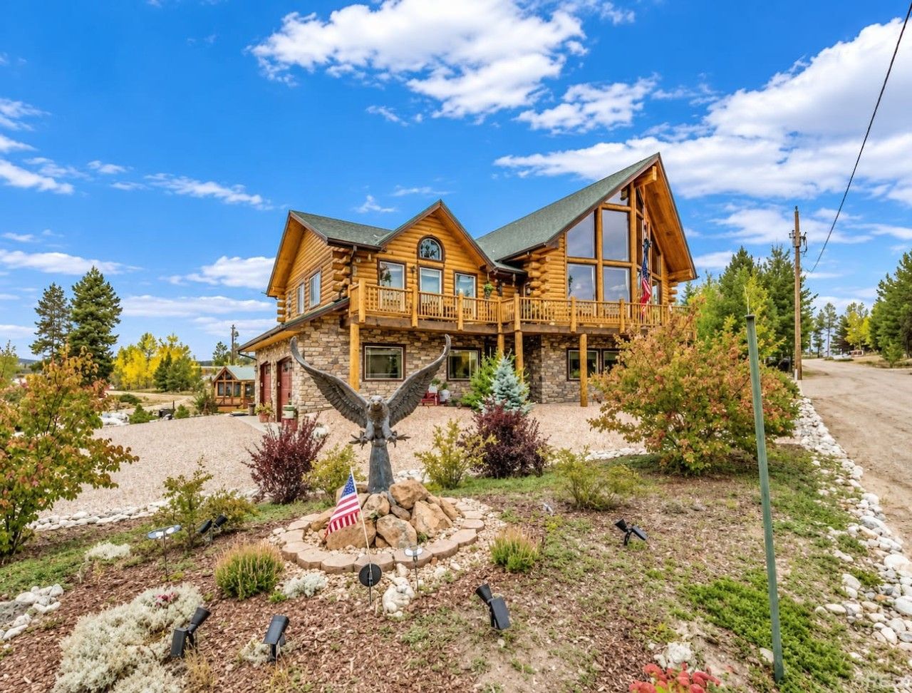 A two-story log cabin with a balcony, surrounded by a landscaped garden with a flag and an eagle statue.