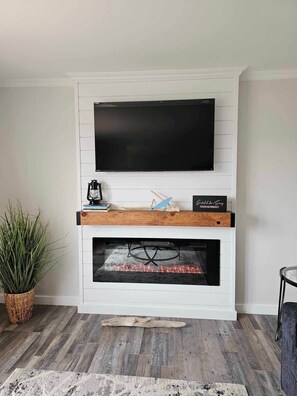 A modern living room with a flat-screen TV mounted above a fireplace, a small table with decorative items, and a potted plant.