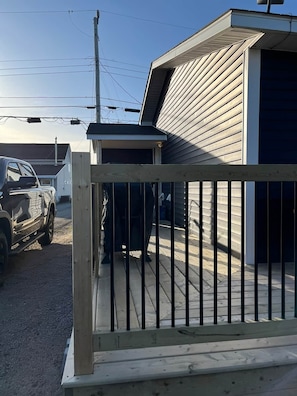 Una terraza de madera con baranda metálica, un vehículo estacionado al fondo y un edificio con techo inclinado.