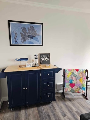 Una habitación con un armario azul oscuro, un mapa enmarcado en la pared, un letrero que dice 'BE OUR guest' y un colcha colorida en una silla.