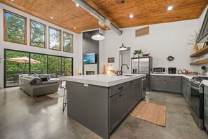 A modern kitchen with a large island, grey cabinetry, stainless steel appliances, and a view to the outdoors. - Secluded Oklahoma Cabin | Hot Tub & Private Acres (Broken Bow)