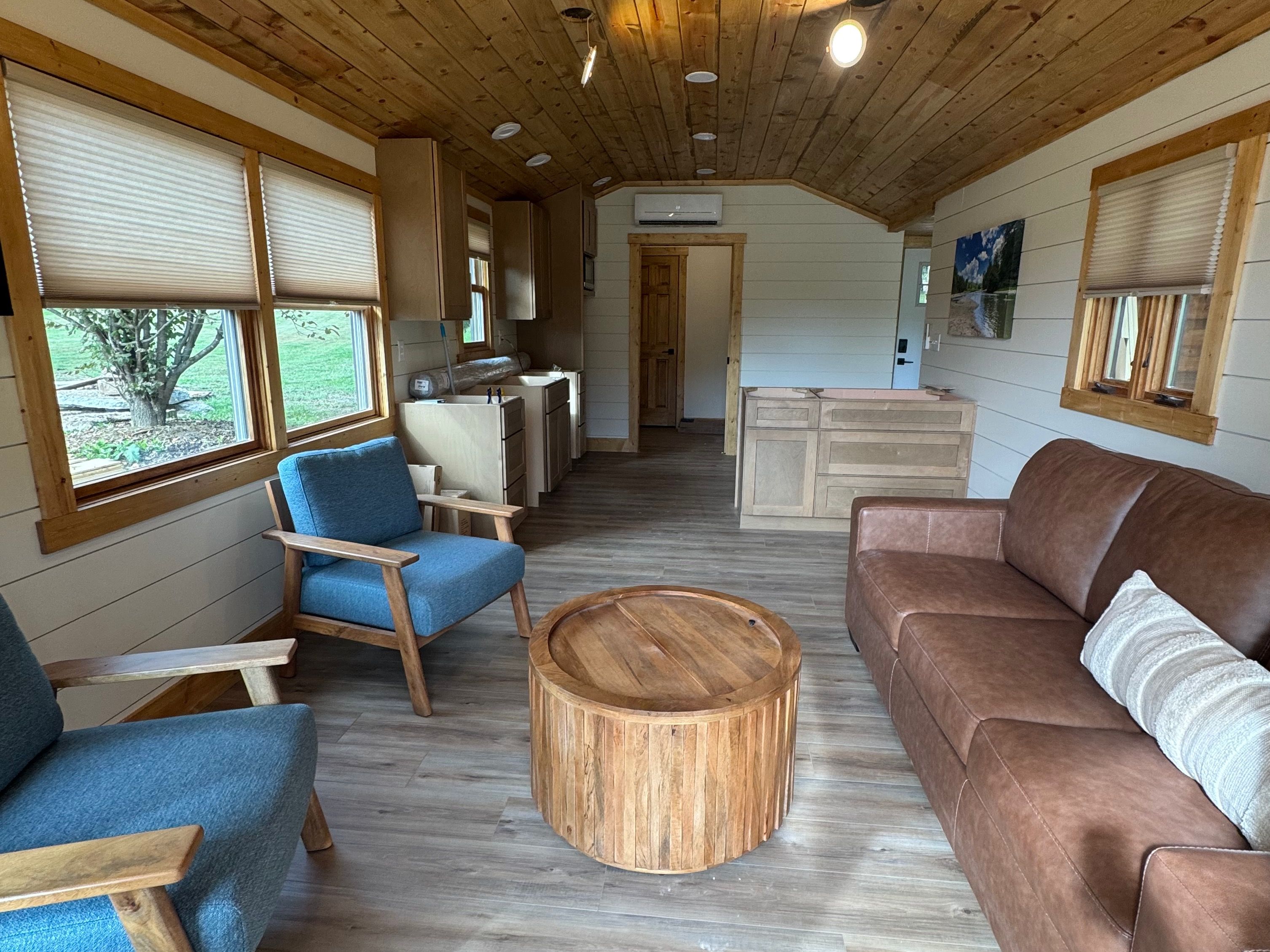 Un intérieur chaleureux avec un plafond en bois, une table basse ronde en bois, un fauteuil bleu, un canapé marron et un coin cuisine équipé d’un évier et de placards.