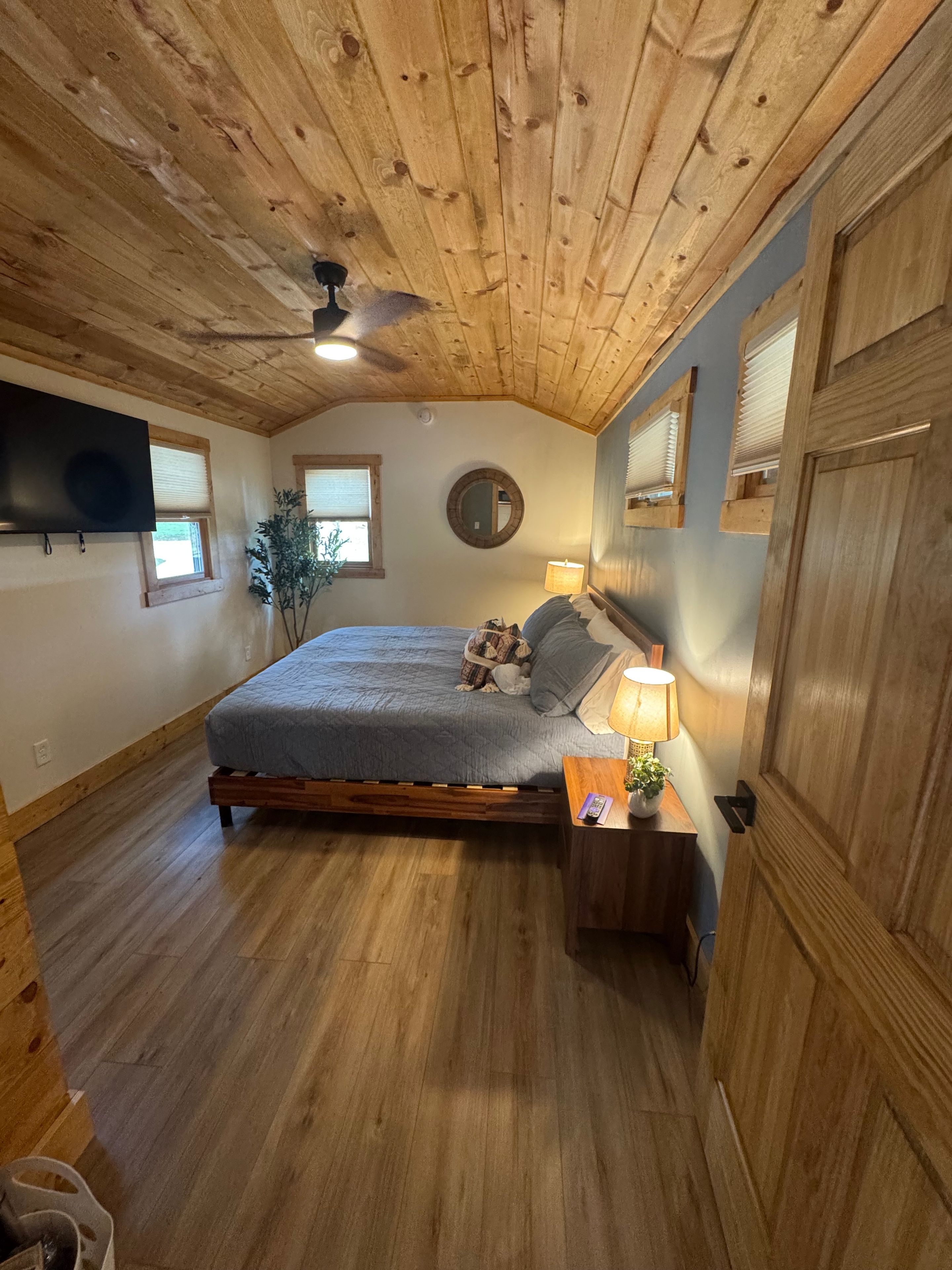 Een slaapkamer met een houten plafond, een bed met een grijze deken, een nachtkastje met een lamp en een aan de muur bevestigde televisie.