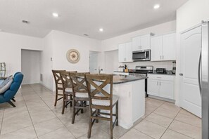 A modern kitchen with white cabinets, a central island, and a dining area with wooden chairs. - Family Getaway | 6BR, Private Pool & Resort (Davenport)