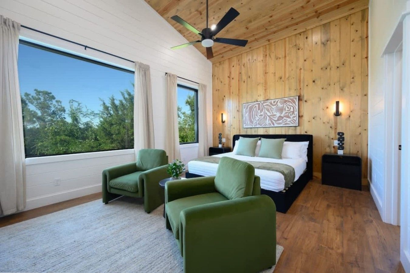 Une chambre à coucher avec un lit, deux fauteuils verts, un ventilateur de plafond, une grande fenêtre et des murs en bois.