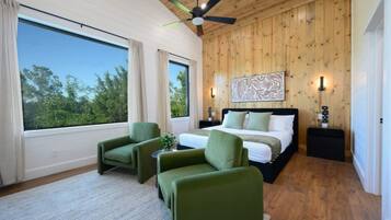 Une chambre à coucher avec un lit, deux fauteuils verts, un ventilateur de plafond, une grande fenêtre et des murs en bois.