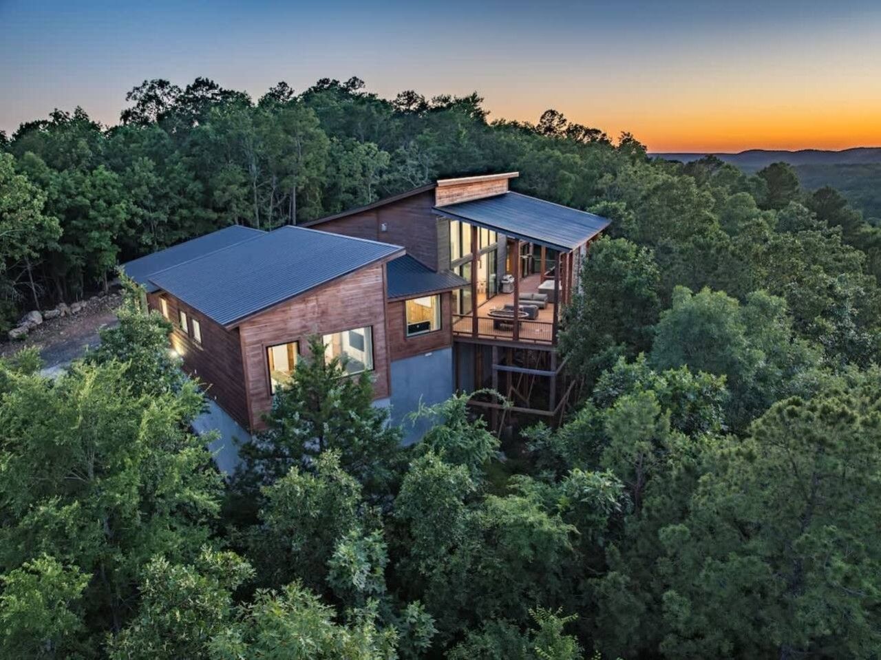 Een modern huis met een groot terras en een balkon op de tweede verdieping, omgeven door dicht bos.