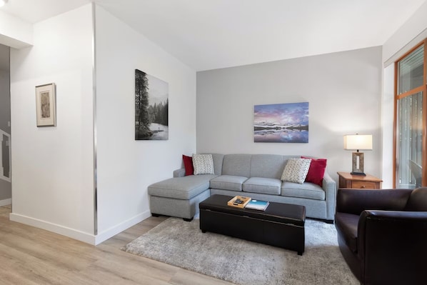 Una sala de estar moderna con un sofá seccional gris, un reposapiés negro, una mesita de madera y una lámpara. Hay dos cuadros enmarcados en la pared.