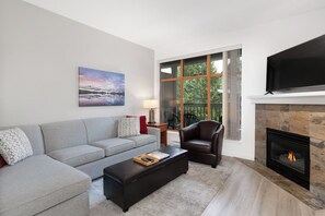 En moderne stue med en grå sofa, en læderlænestol, et fladskærms-tv, en pejs og et stort vindue med udsigt til grønne områder.