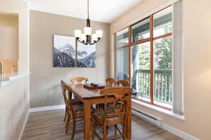 Un coin repas avec une table et des chaises en bois, un lustre, une grande fenêtre donnant sur des arbres et un tableau accroché au mur.