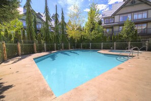 Ein rechteckiger Pool mit grüner Umzäunung und einer Leiter, umgeben von Bäumen und einem Gebäude mit vielen Fenstern und Balkonen.