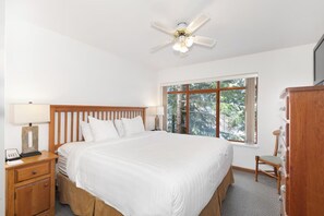 Ein Schlafzimmer mit einem großen Bett, einem hölzernen Kopfteil, einem Nachttisch mit Lampe, einer Kommode und einem Fenster mit Blick auf Bäume.