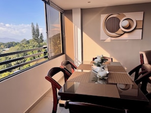 A dining area with a table set for two, chairs, a vase with flowers, and a view of greenery and mountains through a large window. - Amazing views, comfy and close to everything (San Marcos)