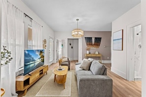 A modern living room with a flat-screen TV, a grey sofa, a wooden coffee table, and a large window with white curtains. - Boho Lake Compound | 2 Homes + Waterfront (Kissimmee)