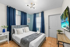 Une chambre à coucher avec un lit, des rideaux, une table de chevet, un téléviseur et une porte.