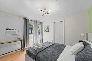 Un dormitorio con cama, mesita de noche, lámpara, lámpara de techo, ventana con cortinas y una puerta.