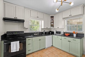 Una cocina con armarios blancos, una estufa negra, un microondas y una ventana con persianas.