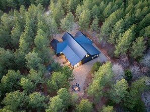 A modern house surrounded by dense forest, with a visible patio area and a fire pit. - Steel Time Cabin | Ping Pong | Jacuzzi | Fire Pit (Broken Bow)