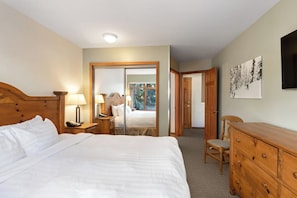 Une chambre avec un grand lit, une tête de lit en bois, des tables de chevet avec des lampes, une commode et un miroir. Il y a une porte qui donne sur une autre pièce et une fenêtre avec vue.