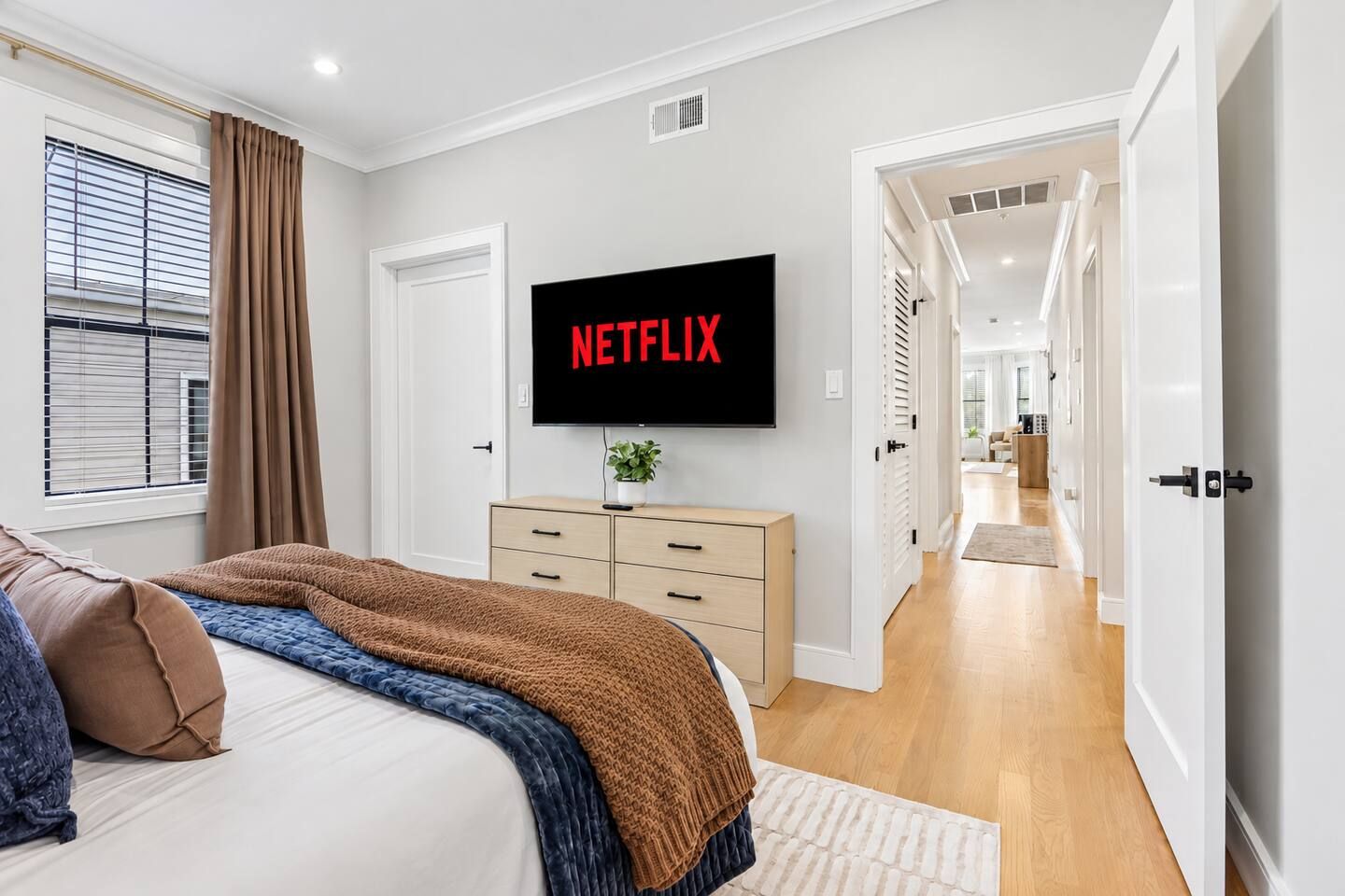 Een slaapkamer met een bed, een aan de muur bevestigde televisie, een dressoir en een raam met gordijnen.
