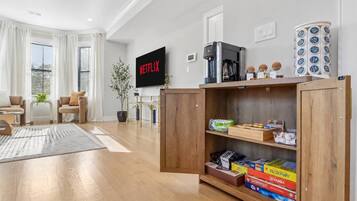 A modern living room with a flat-screen TV, wooden shelves with board games, and a coffee machine.