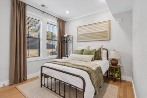 A bedroom with a bed, a bedside table with a lamp, a window with curtains, and a wall-mounted artwork. - 4BR Boston Stay Near Orange Line (Boston)