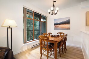 Un área de comedor con una mesa y sillas de madera, una lámpara de pie, un ventanal con vistas a árboles y un cuadro en la pared.