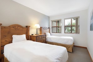 Un dormitorio con dos camas, cabeceros de madera, una mesita de noche con lámpara y dos ventanas.
