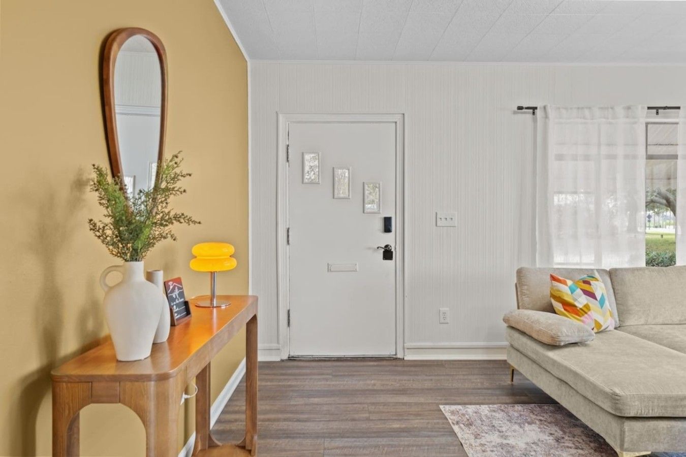 A living room with a sofa, a wooden console table with a vase and a lamp, a white door, and a window with curtains.