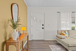 A living room with a sofa, a wooden console table with a vase and a lamp, a white door, and a window with curtains. - Boho Lakeside Retreat | Walk to Lake Toho (Kissimmee)
