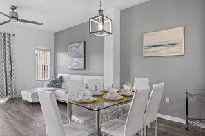 A modern dining area with a glass table, white chairs, and a ceiling fan. - Lake Berkley Retreat | Pool • Fishing • Clubhouse (Kissimmee)