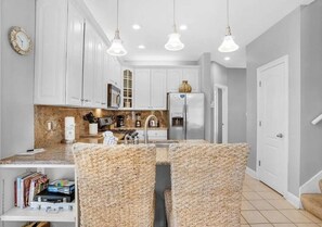 A kitchen with white cabinets, a built-in oven, and a refrigerator. There are wicker chairs and a dining table with books. - Destin Family Retreat | Private Pool + Beach (Destin)