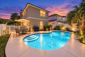 A swimming pool with a curved edge, surrounded by a white fence, and a patio area with chairs and a table. - Destin Family Retreat | Private Pool + Beach (Destin)